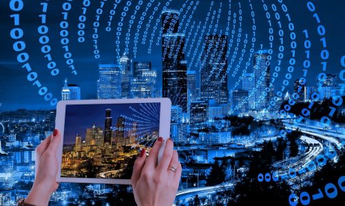 Person mit Tablet, die die Skyline einer Stadt fotografiert über der ein Binary Code schwebt.