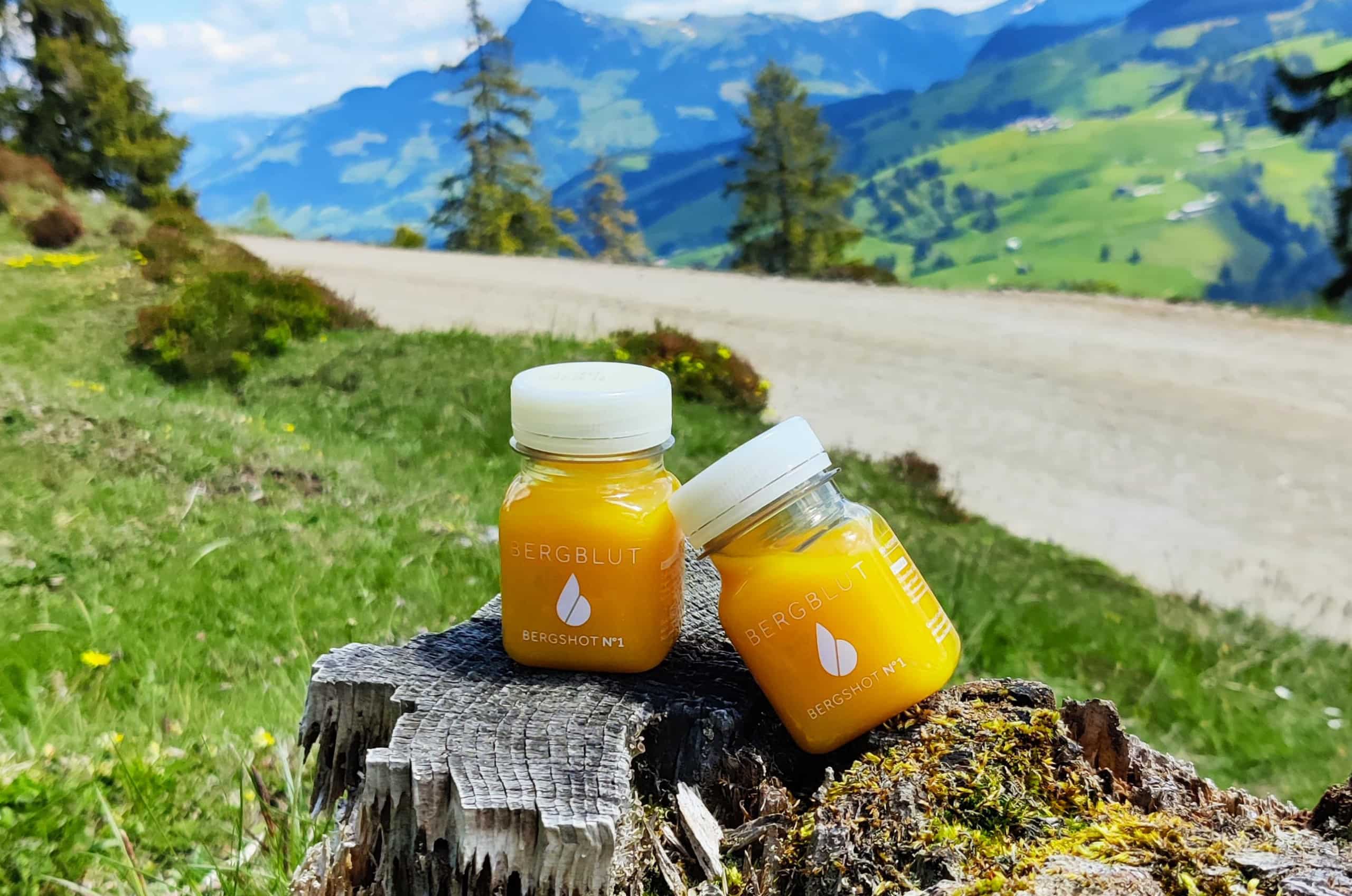 Zwei orangefarbene Säfte stehen auf einem Baumstumpf an einem Wanderweg in den Bergen. Nachhaltiges Marketing bei Bergblut.